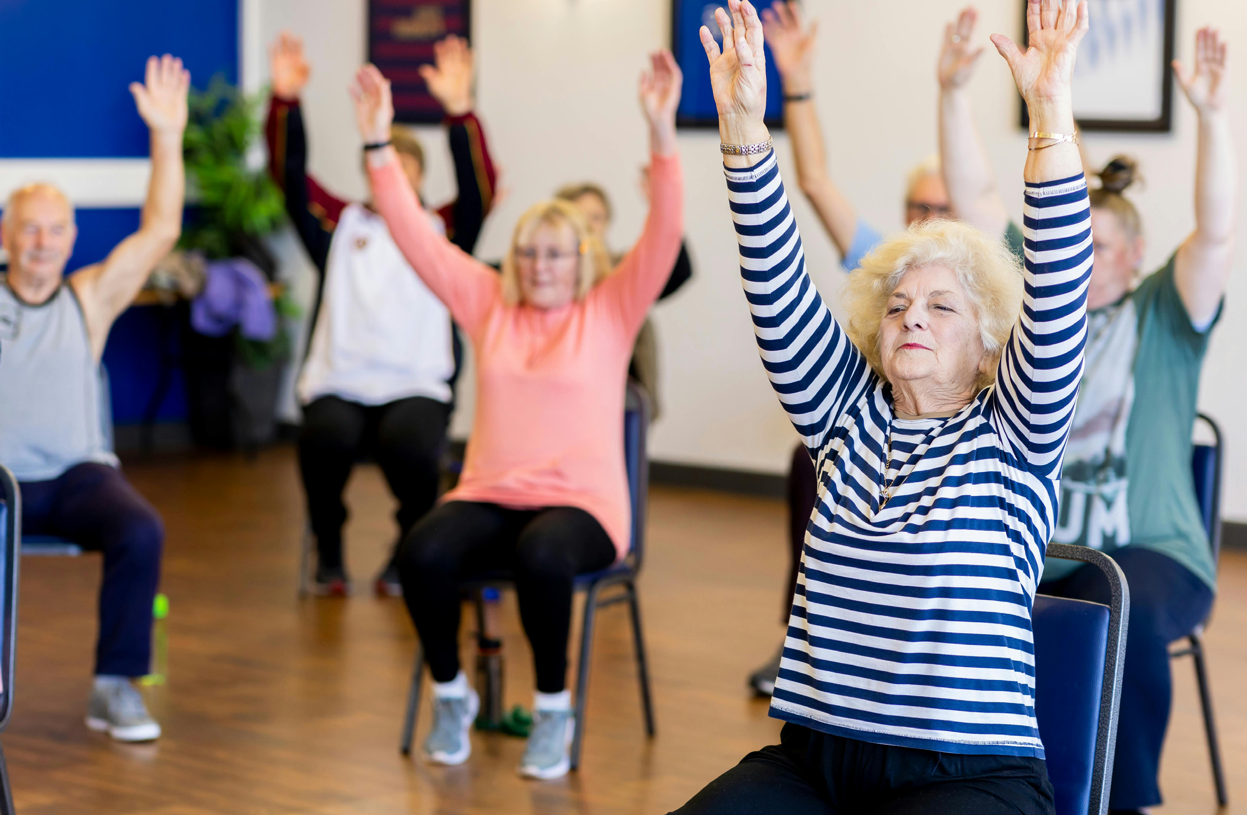 A class of seven older adults in chairs stretching their arms upwards.